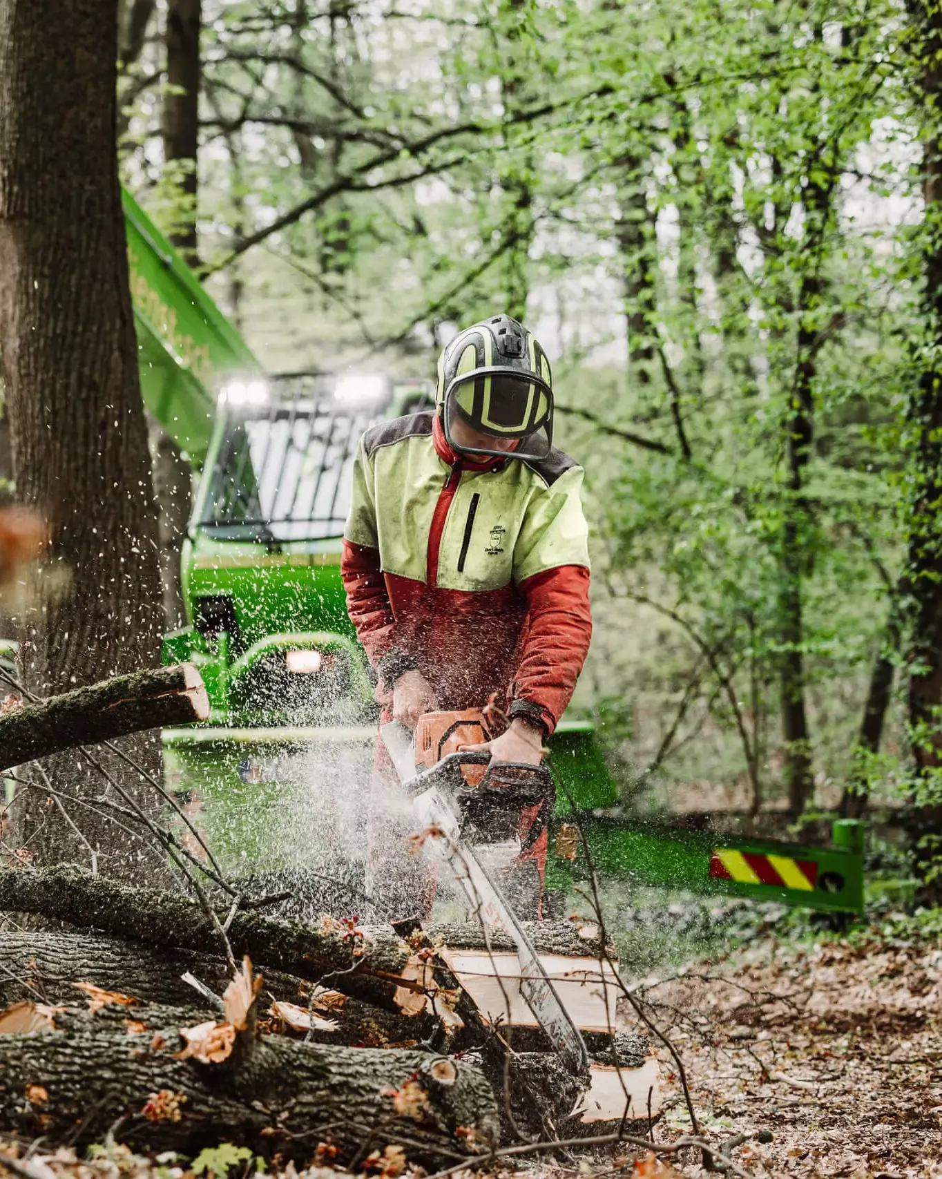 Eine Person mit Schutzausrüstung sägt mit einer Kettensäge Stämme in einem Wald. Im Hintergrund ist ein grünes Forstfahrzeug zu sehen, und Sägespäne fliegen durch die Luft.