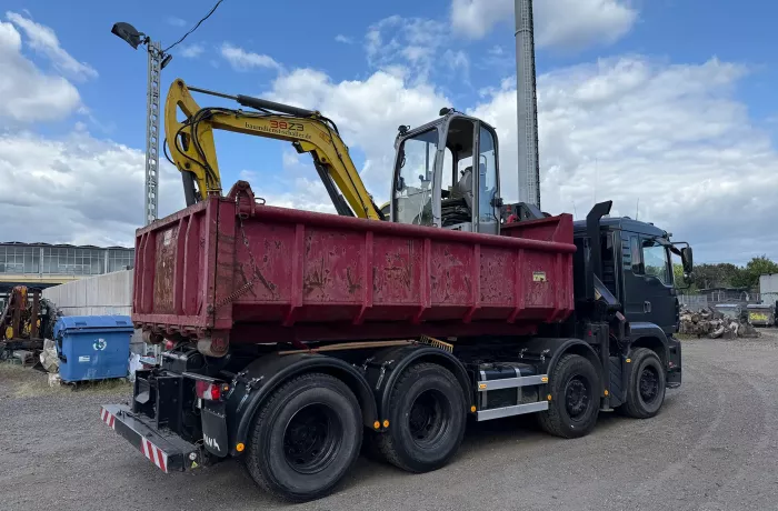 Ein schwarzer Lkw mit einem roten Container auf offener Ladefläche transportiert einen kleinen gelben Bagger. Der Lkw ist auf einem Kiesplatz geparkt, mit Industriegebäuden, Geräten und einem blauen Himmel mit Wolken im Hintergrund.